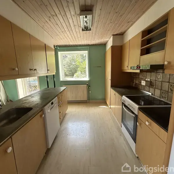 Køkken med træloft, brunlige skabe og sort bordplade, har ovn, vask og opvaskemaskine. Et vindue kaster lys ind, og en køleskab står til højre. Vægge har grøn nuance.