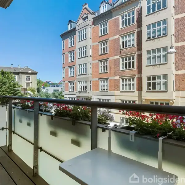 En balkon med træterrasse og blomsterkasser langs rækværket, i rolig bymiljø omgivet af klassiske murstensbygninger. Klar blå himmel i baggrunden.