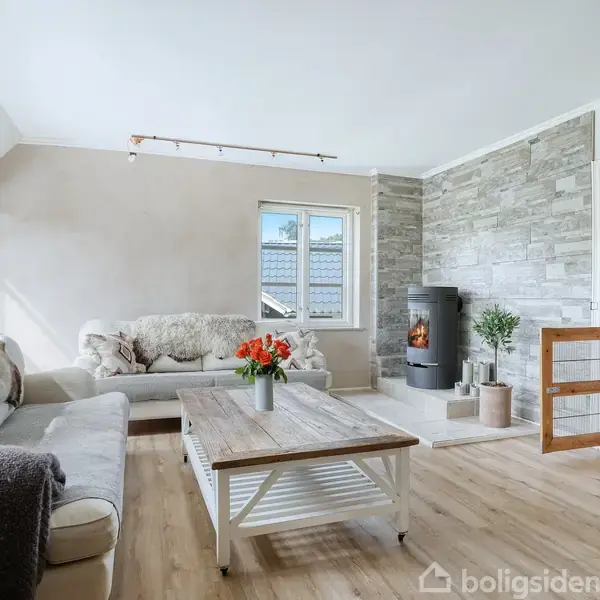 Stue med to lyse sofaer arrangeret rundt om et træbord, der har en vase med blomster. En brændeovn står ved væggen i et lyst og indbydende rum.