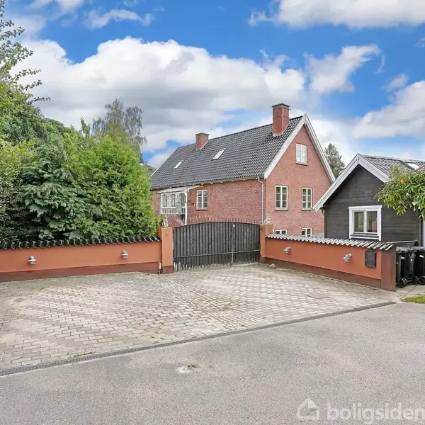 En rød murstensbygning står stille bag en sort port omkranset af høje hække. Foran huset er en brolagt indkørsel omgivet af lave mure, mens en blå himmel hænger over.