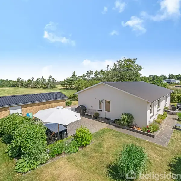 Et hvidt hus med en mørk tag udfolder sig i en velplejet have. En terrasse med parasol er synlig, omgivet af grønne buske og græsplæner. Skov i baggrunden.
