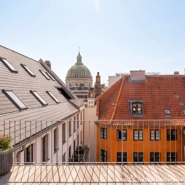 En balkon med udsigt over byens hustage. Brostensbelagte tage omgiver en gård. I baggrunden ses en stor kuppel, der tilhører en prominent bygning, mod en klar blå himmel.