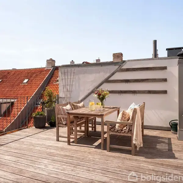 Træterrasse med bord og stole arrangeret til måltid. Omfattende blomsterdekorationer på bordet. Sollys skinner stærkt på scenen. Baggrunden viser hustage og klare himmel.