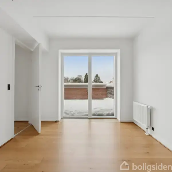 Et lyst, tomt rum med trægulv har en åben dør til venstre og en stor glasdør, der fører ud til en altan. Udenfor ses snedækkede træer.