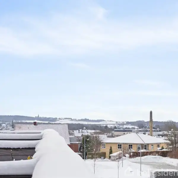 En snedækket by ses i horisonten under en klar blå himmel. Venstre side viser en del af en bygning med en balkon.