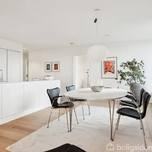 Spisebord med sorte stole er placeret på et hvidt tæppe i et lyst, moderne køkken-alrum. Bagved ses et hvidt køkkenmodul med en indbygget ovn og dekorationer på væggene.