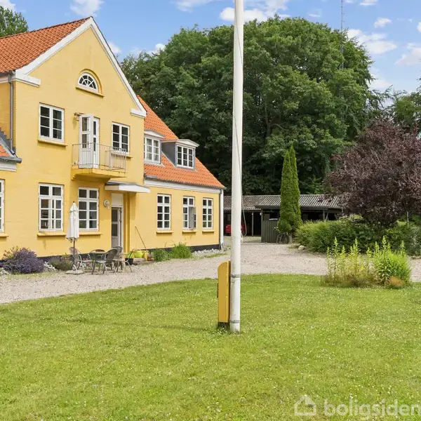En gul to-etagers bygning med rødt tegltag og flere vinduer. Foran står en flagstang på en grøn plæne omgivet af grus. Omkring er der træer og en carport i baggrunden.