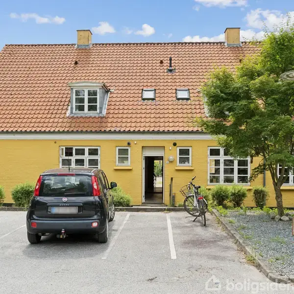 Gul bygning med rødt tegltag. En sort bil og cykel står parkeret foran på en asfalteret indkørsel. Træer og buske omkranser stedet for en rolig atmosfære.