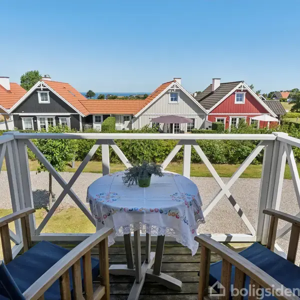 To træstole står omkring et bord med dug på en altan. Bordet har en lille dekorativ plante. I baggrunden ses farverige huse og en klar blå himmel.