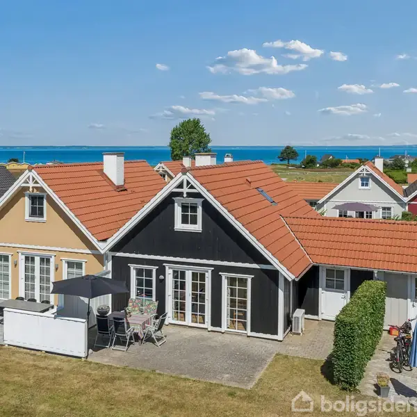 Et rækkehus i sort og hvid står stille i en have med terrassemøbler og parasol, beliggende nær kysten. I baggrunden ses flere huse og havet under en blå himmel.