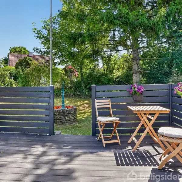 Terrasse med to stole og et bord står på et trægulv. Stolene har puder, og blomster pryder rækværket. Baggrunden består af en have med træer og buske.