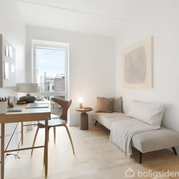 Et lyst rum med en skrivebordsplads til venstre og en grå sofa til højre. En vinduesramme viser en udendørs scene. Væggene er dekoreret med minimalistisk kunst.