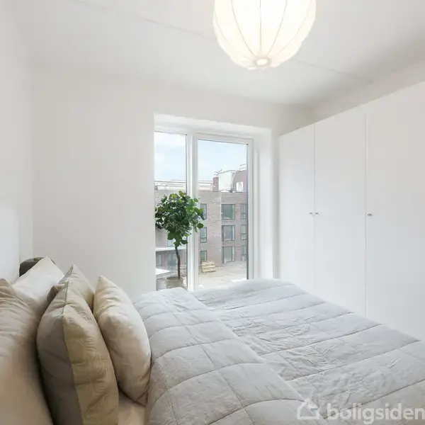 Soveværelse med en stor dobbeltseng dækket af en grå dyne, står ved siden af et vindue med udsigt til en bygning. Skab og en lille plante ses ved siden af.