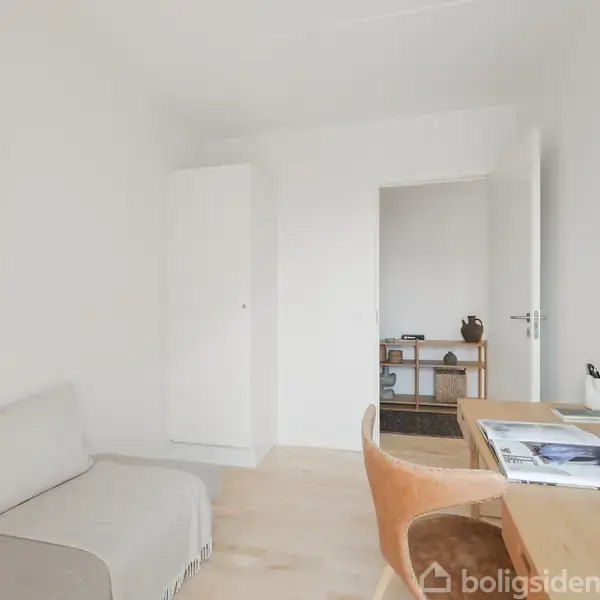 Sofa står i et lyst rum med en beige plaid. Ved siden af er der et skrivebord med bøger og en stol. Vægge er dekoreret med kunst.