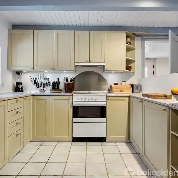 Et lyst køkken med beige skabe og hvide flisegulve. En komfur står centralt mellem skabe, omgivet af køkkenredskaber og en brødkasse på bordpladerne. Vinduer bringer naturligt lys ind.