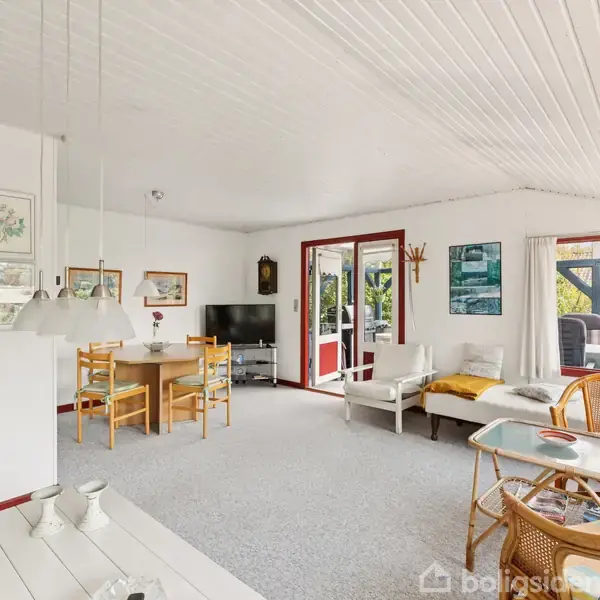 Stue med sofa og stole omkring et sofabord, spisestue i baggrunden med træbord og stole, fladskærms-TV i hjørnet, store vinduer med udsigt til terrasse og have.