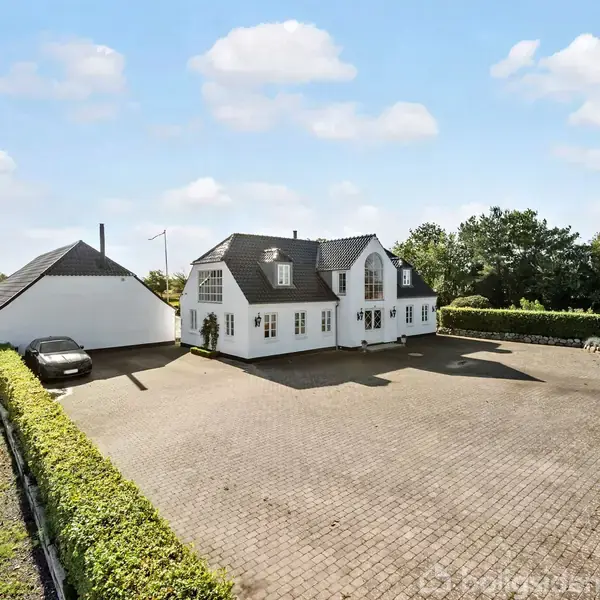 Et hvidt hus med sorte tage står stille på en stor flisebelagt gårdsplads omgivet af hæk. En bil er parkeret ved en mindre bygning. Skoven og markerne omslutter ejendommen.
