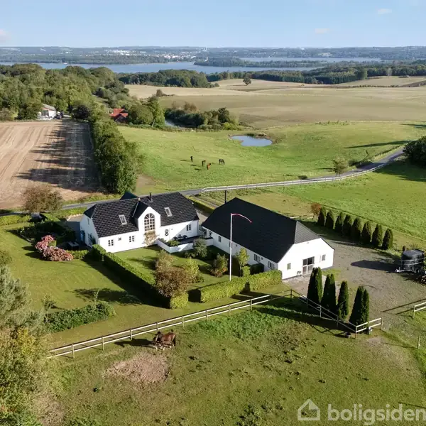 En hvid gård med sort tag ligger i et frodigt landskab med marker og skove. Der er en indhegning med en hest foran huset og marker omkring.