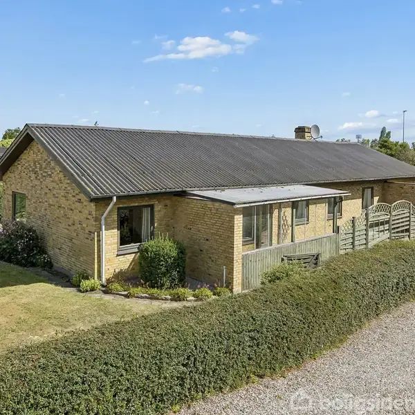 En gulstensvilla med sort tag står stille i en haveomgivelser. Huset er omkranset af en velplejet hæk og græsplæne, under en klar blå himmel.