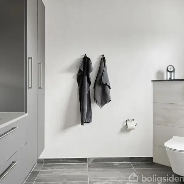 Gråt badeværelse med moderne skabsindretning og håndklæder på væggen. Toilet til højre med plante, tandbørsteholder og toiletrulleholder. Flisegulv og lys farvepalet.
