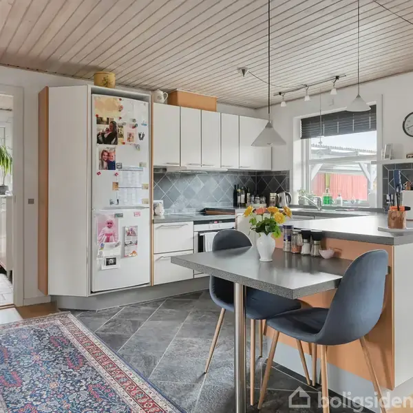Køkken med hvidt skab som har fotos på siden og en bordplade med blomster i en vase. To stole ved bordet, flisegulv, tæppe og vindue med udsigt til en rød bygning.