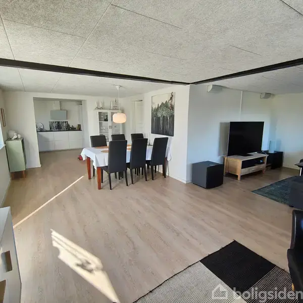 Spisestue med sort stole omkring hvidt dækket bord under pendellampe; til højre, stueområde med tv og sort sofa; lyst trægulv og store vinduer til venstre.