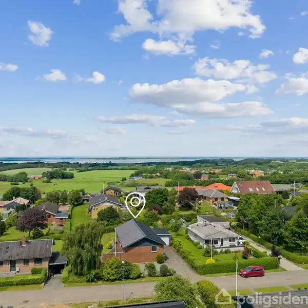 Hus med grå tag markeret med en cirkel ligger i en forstad. Omgivet af andre boliger, grønne træer og en vej. Under en blå himmel med spredte skyer.