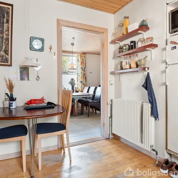 Et køkken med et rundt bord og to stole. Bordet har papir og en krukke med redskaber. Der er hylder med glas og skåle samt en mikrobølgeovn oven på køleskabet.
