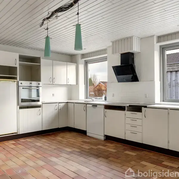 Køkken med hvide skabe og indbygget ovn. Lys strømmer ind gennem to vinduer. To hængelamper hænger fra loftet. Gulvet er af terracotta-fliser.