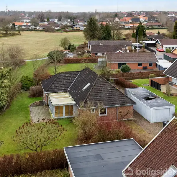 Et parcelhus med en tagterrasse står omgivet af en have med grønne træer. Nabohuse og en stor mark udfylder den landlige baggrund.