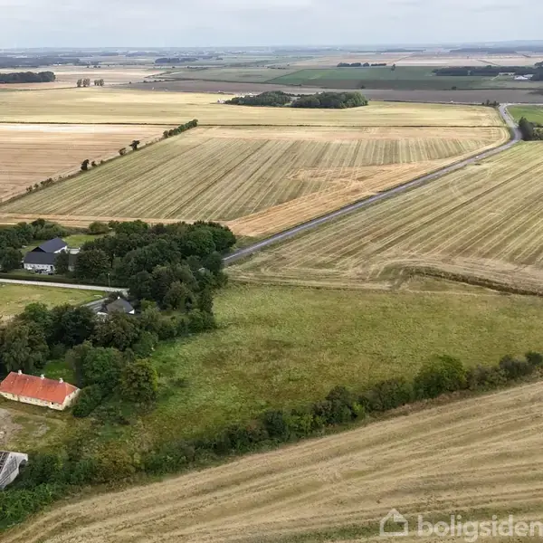 Luftfoto af åbne marker med en vej, der løber gennem landskabet. Et par huse ses omgivet af træer i forgrunden, bagved strækker markerne sig mod horisonten.