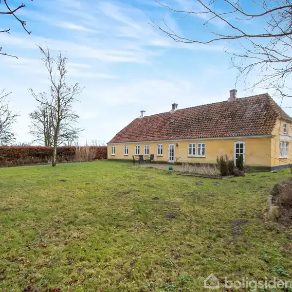 Gult hus står stille i en åben have med træer og hæk i baggrunden under en blå himmel.