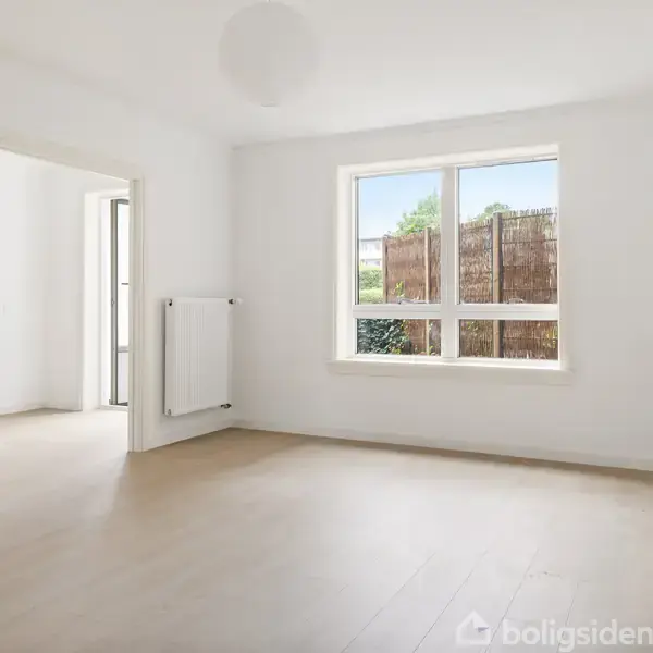 Et lyst værelse med et stort vindue slipper dagslys ind. En radiator er fastgjort til væggen. Rummet fører til en anden sektion med lignende gulv og vægge.