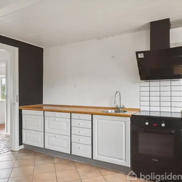 Køkken med hvide skabe og brun bordplade. En sort ovn og emhætte er placeret ved fliser. Rummet er lyst med flisegulv, og der er en dør til et tilstødende område.