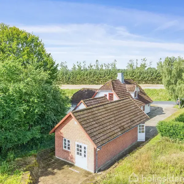 Rødt murstenshus med mørkt tegltag omgivet af træer og græsplæne. Solbeskinnet gårdsplads foran huset, som er placeret i et landligt område med hække og åben blå himmel.