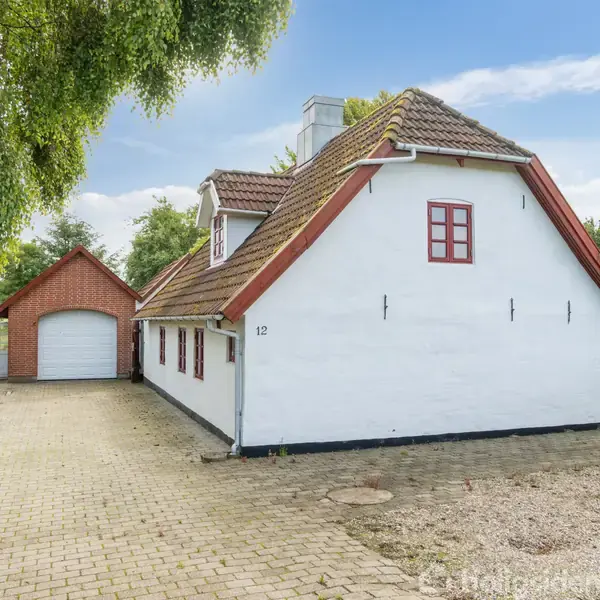 Hvidt hus med rødt tag og små vinduer står ved siden af en garage. Omkringliggende have og brolagt indkørsel. Nummeret "12" er på husets facade.