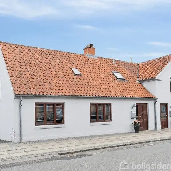 Hvidmalet hus med rødt tegltag står stille ved en vej, omgivet af andre huse. Taget har to små vinduer, og ved indgangen står en plantepotte.