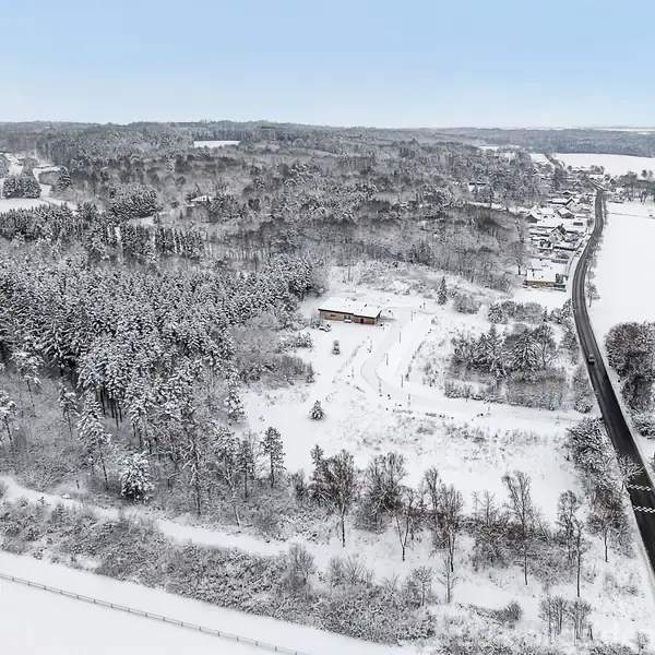 Sneklædt landskab ses fra luften, med en træbygning midt i en skov. En vej strækker sig gennem det naturskønne, vinterlige område til en lille bebyggelse i baggrunden.