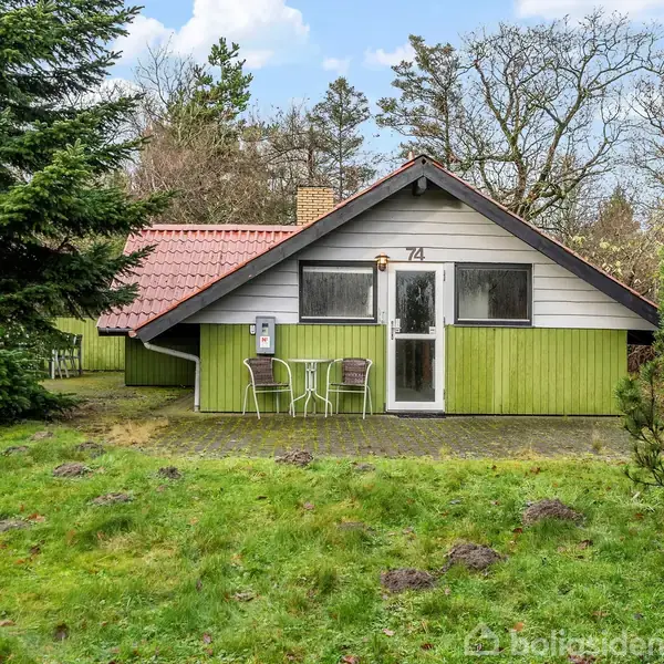 Et lille hus med et rødt tag og grøn facade har to stole foran døren, omgivet af en frodig have og træer. Nummer 74 ses ved indgangen.