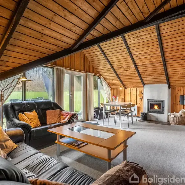 Et træbeklædt opholdsrum har en sort lædersofa med puder foran en glasbord, pejs i baggrunden, med store vinduer der afslører en grøn græsplæne udenfor.