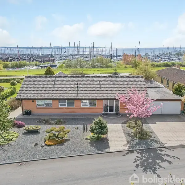En etplanshus med gråt tag og orange facade står i en have med sten og træer. Det ligger nær en marina med mange både, omgivet af græsarealer og andre huse.