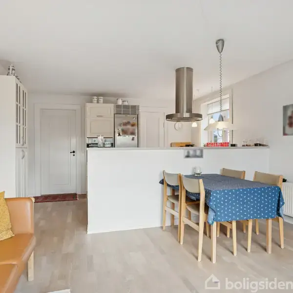 En stue med spisebord og fire stole dækket med en blå dug, placeret ved en halv væg adskiller køkkenet med hvide skabe. Orange sofa til venstre, hylde på væggen.