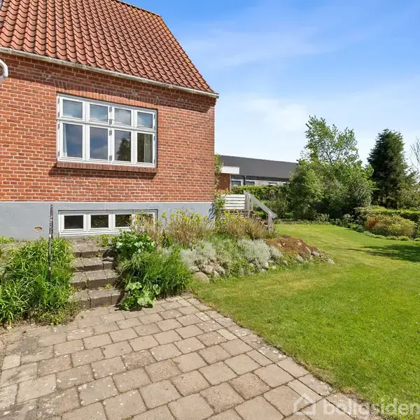 Rødt murstenshus med et rødt tegltag. Huset har en stor vinduesrude. Flisebelagt terrasse fører til en frodig have med græsplæne og buske i solrigt vejr.