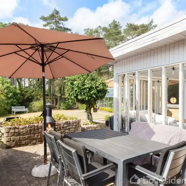 Terrassemøbler med parasol står på flisebelagt terrasse, mens det tilstødende hus med store vinduer har udsigt til en grøn have og skov i baggrunden. Et skilt viser "Voresbo".