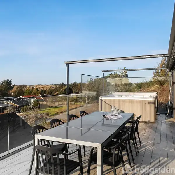 Langt havebord med sorte stole står på en stor træterrasse. Boblebad i baggrunden. Omgivet af et glasværn, udsigt over et boligområde under en blå himmel.