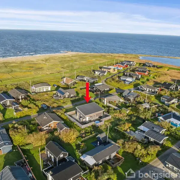 En kystnær landsby med spredte huse omgivet af grønne områder, peget ud af en rød pil. I baggrunden ses havet og stranden.