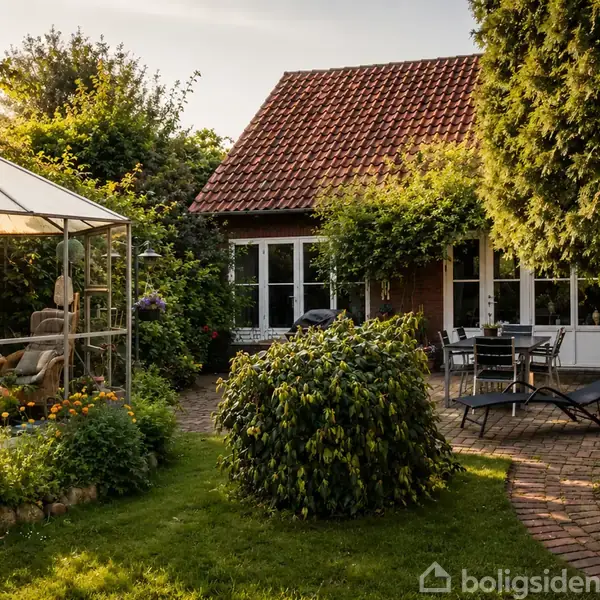 Et hyggeligt haveområde med et drivhus til venstre, omgivet af blomster og planter. Foran et rødt tegltagshus står der et bord og stole på en stenlagt terrasse.