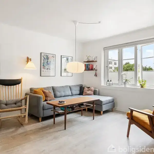 Gråt sofaarrangement står i en lys stue, flankeret af en træstol til venstre og orange polstret stol til højre. Væggene har kunstværker. Store vinduer slipper lys ind.