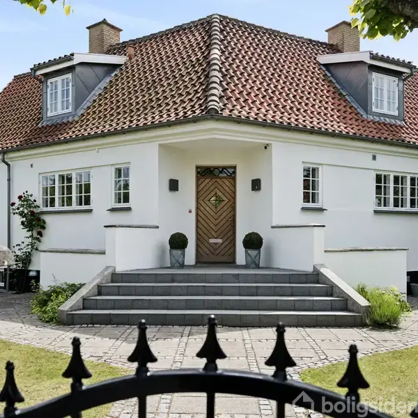 Et hvidt, klassisk hus med rødt tegltag og to kviste. Huset har en central trappeindgang med to små buske på hver side, omgivet af en velplejet have.
