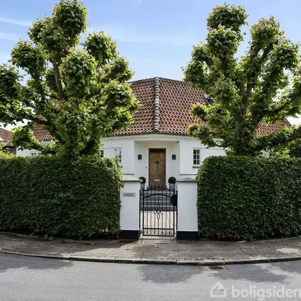 Hvid villa med rødt tegltag ses bag en høj hæk. Foran huset er der en sort låge og to formklippede træer. Beliggende på en rolig villavej.